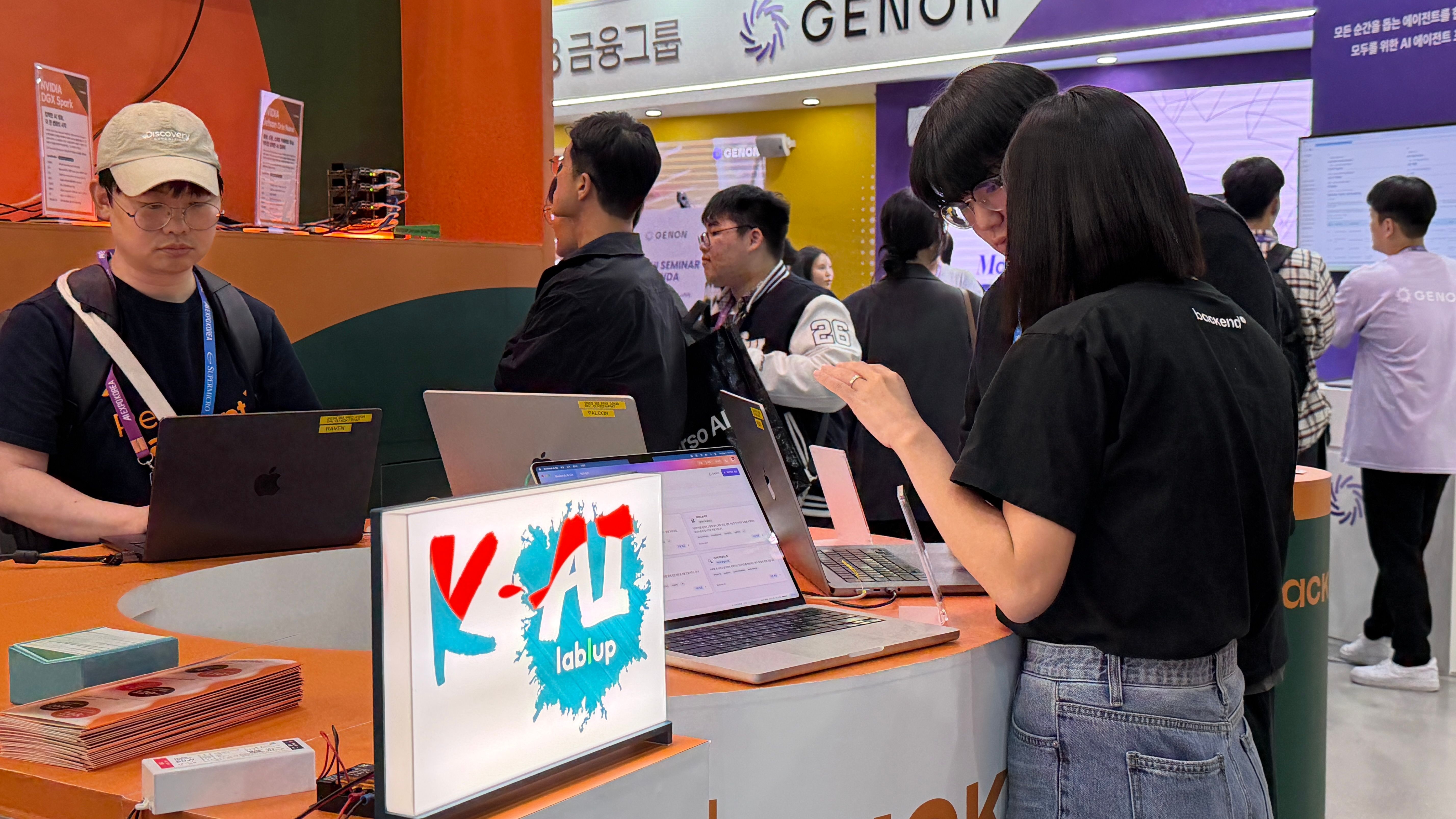 Three MacBooks lined up at the booth next to a K-AI lablup display, set up for AI:GO demos.
